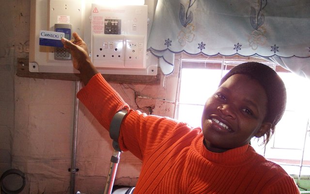 â€˜n Vreugdevolle gestremde Bukelwa Mbuzweni by haar elektrisiteitstoestel vir elektrisiteit in haar woning